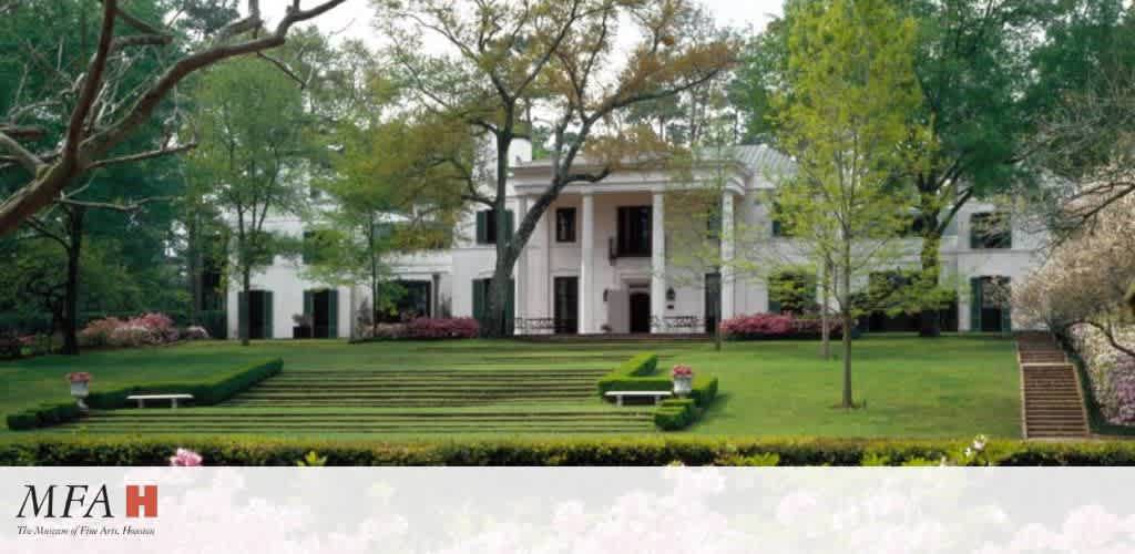 A grand white mansion is set amidst lush greenery. The front yard features neatly manicured lawns with tiered steps leading to the entrance. Tall trees surround the area, providing shade and elegance. Pink and red flowers add color to the landscape. The setting evokes a sense of tranquility and refinement.
