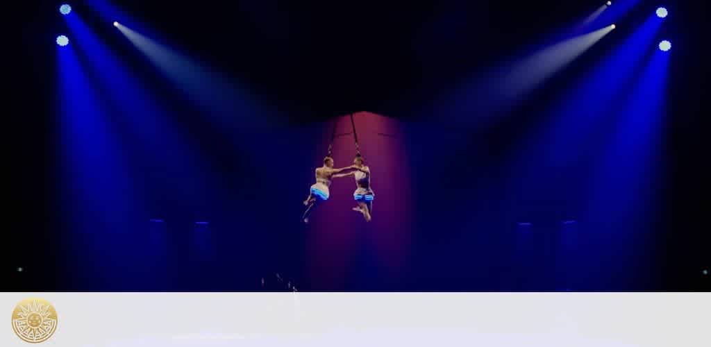 Two trapeze artists in mid-air, performing under blue and purple spotlights. The performers, dressed in white and blue outfits, are suspended in a dark theater setting, showcasing agility and grace. The image captures a moment of dynamic motion and balance, creating an atmosphere of excitement and wonder suitable for an acrobatic performance.