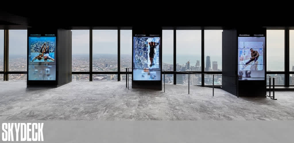 Spacious observation area at Skydeck showcasing tall glass windows with panoramic city views. Three large digital screens display adventurous activities. The floor is cement, and a barrier runs along the windows for safety. The word Skydeck is visible in the bottom left corner. The scene suggests an interactive and modern viewing experience.