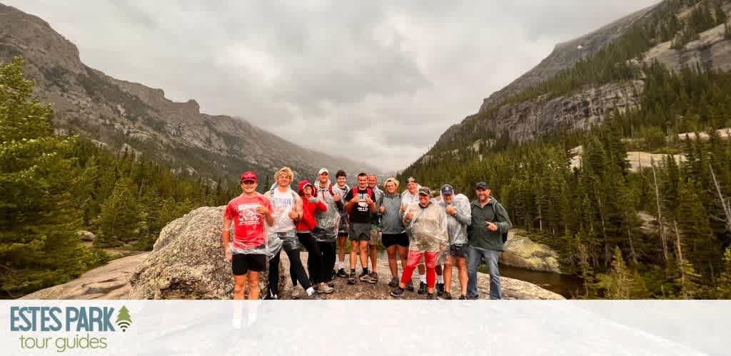 A group of people stands on a large rock in a scenic mountain setting. They are dressed in casual outdoor clothing, some with rain gear, and are smiling at the camera. Lush green pine trees surround them, and towering mountains rise in the background under a cloudy sky. The words Estes Park Tour Guides are at the bottom left.