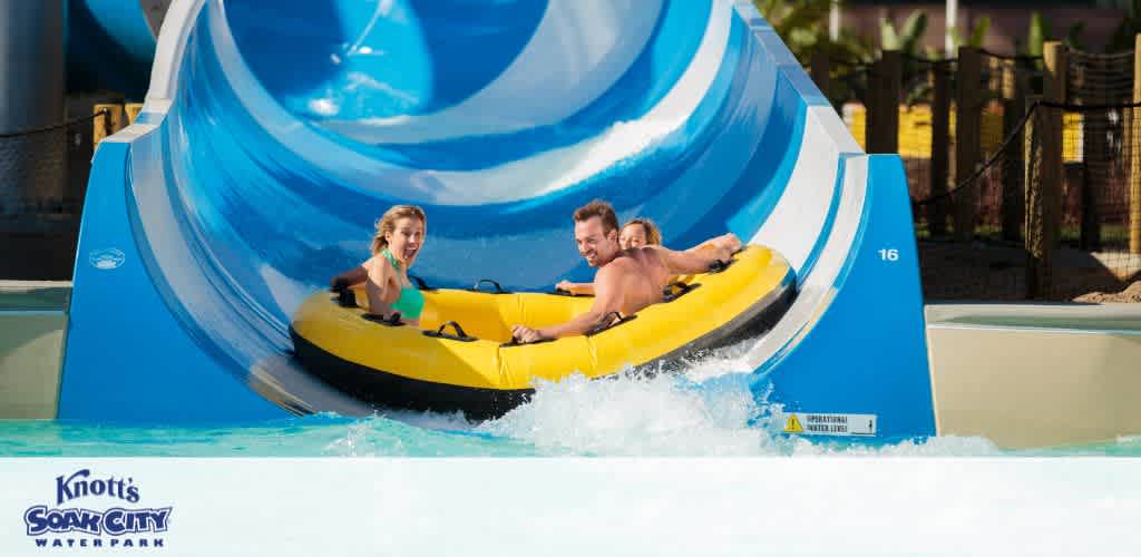 This image captures the thrill of a water slide experience at Knott's Soak City Water Park. Two riders, a woman in the front and a man in the back, are shown seated in a bright yellow inflatable raft as they emerge from a large blue water slide with a white and blue spiral design. They appear to be laughing and enjoying the ride, with water splashing around them as they hit the bottom of the slide, indicating a fun and exhilarating moment. The woman in the front of the raft has her arms raised in excitement while the man holds onto the raft handles. The number 16 is visible on the side of the water slide exit, hinting at multiple slides available in the park. 

Remember, when you book your fun-filled adventure with GreatWorkPerks.com, you're guaranteed to enjoy the lowest prices and great savings on tickets!