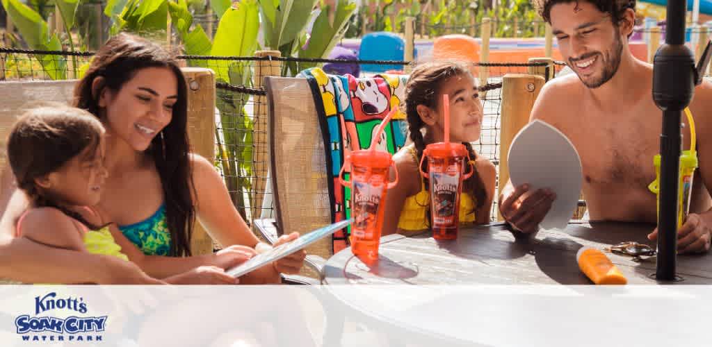 Image Description: The photo captures a cheerful family moment at Knott's Soak City Waterpark. To the left, a smiling woman with dark hair in a green floral swimsuit is seated next to a young girl with dark hair; they are both looking at a book or menu together. At the center of the image, another young girl with dark hair and wearing a yellow swim shirt is leaning on a table, with the sunlight enhancing her bright expression as she gazes adoringly at a man to her right. The man, who has a beard and is shirtless, is smiling back at the girl while holding up a white, circular object, which could be a plate or dish. Behind them, colorful life vests hang on a bamboo fence, and tropical foliage is visible, adding to the summery atmosphere of the setting. On the table are two branded orange water bottles with Knott's Soak City logos.

End with promotional sentence: Don't miss out on family fun—visit GreatWorkPerks.com for the lowest prices and exclusive discounts on tickets to memorable experiences like a day at Knott's Soak City Waterpark!