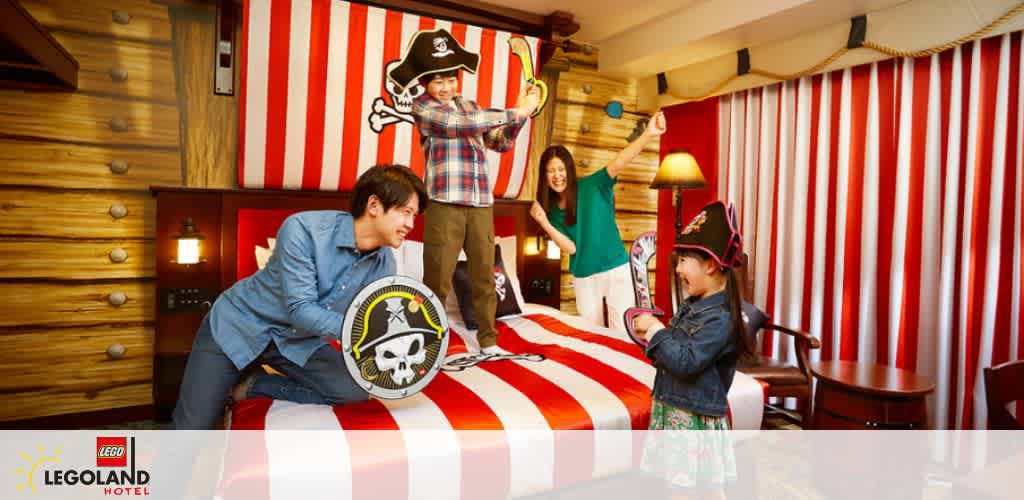 A pirate-themed room at LEGOLAND Hotel with a family enjoying playtime. A child wearing a pirate hat stands on the bed with a sword. Another child holds a shield. Two adults in the background cheer and play along with joyful expressions. The room features bright red and white striped walls and wooden decor.