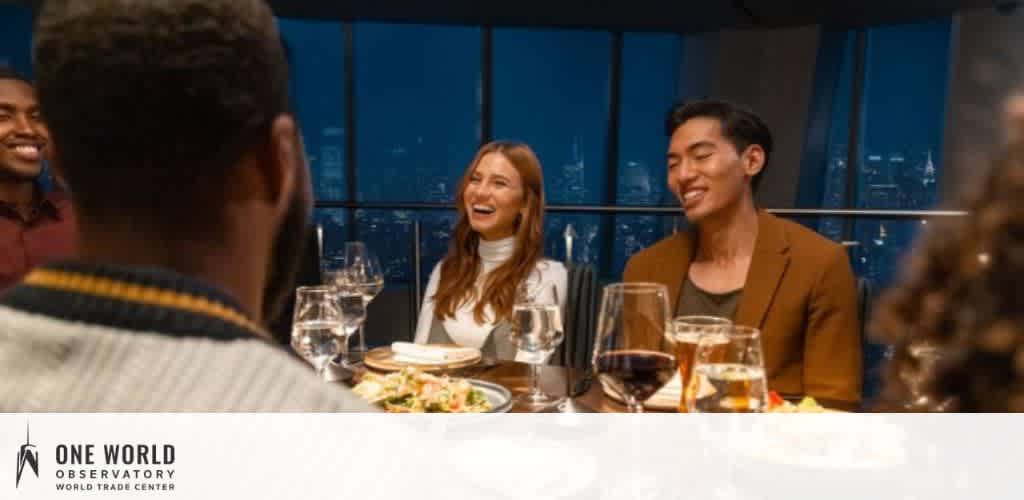 A group of people is enjoying a meal at a dimly lit restaurant. In the foreground, a person serves food to a smiling woman. In the background, other guests engage in conversation. The logo of One World Observatory is visible in the bottom left, indicating the high-altitude dining experience at the World Trade Center.