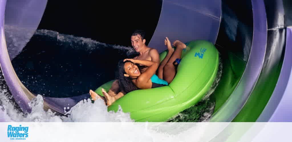 Image shows two people with delighted expressions sliding down a water chute in a shared green inflatable ring. Water splashes around them, and the Raging Waters Los Angeles logo is visible in the corner.