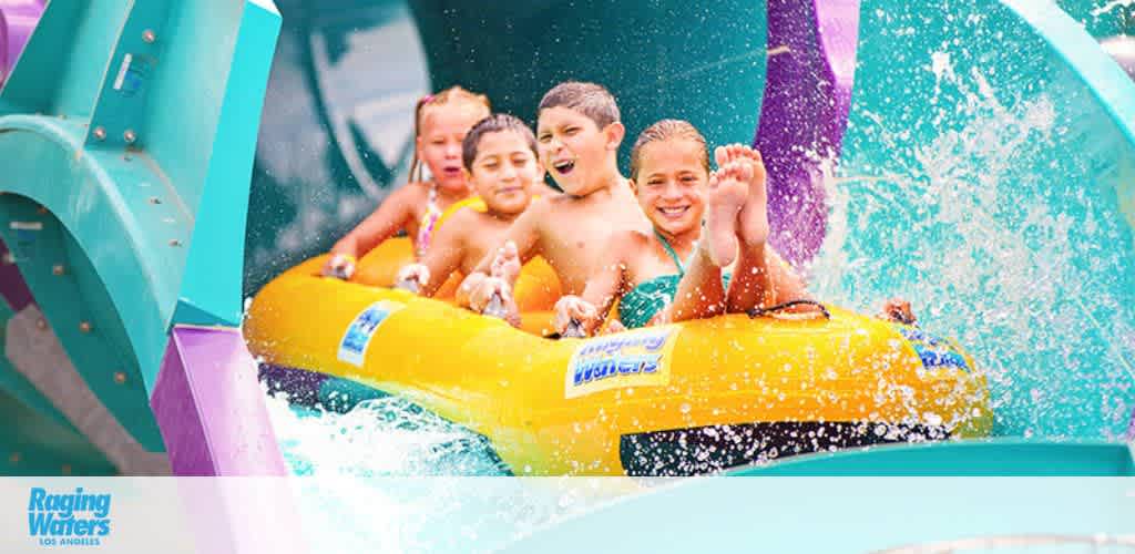 Image displays a group of joyful children sliding down a colorful water slide on a yellow raft at Raging Waters, Los Angeles. Splashing water and vibrant colors contribute to the lively atmosphere of the scene.