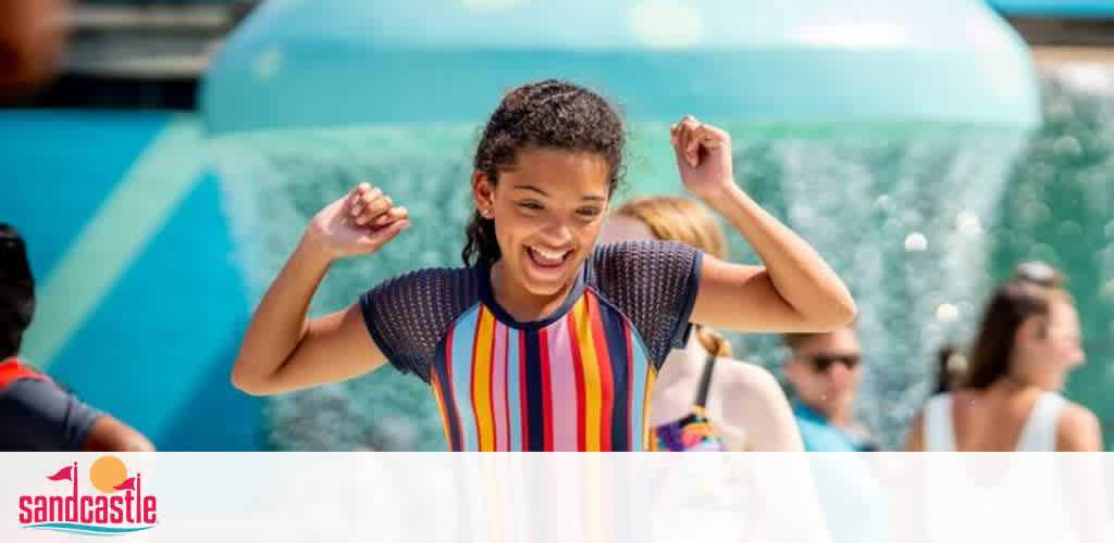 A joyful young girl with her arms raised in excitement stands in front of a water ride at Sandcastle water park. She is smiling broadly, wearing a colorful striped swim shirt, enjoying a sunny day out. People and water splashes can be seen in the background.