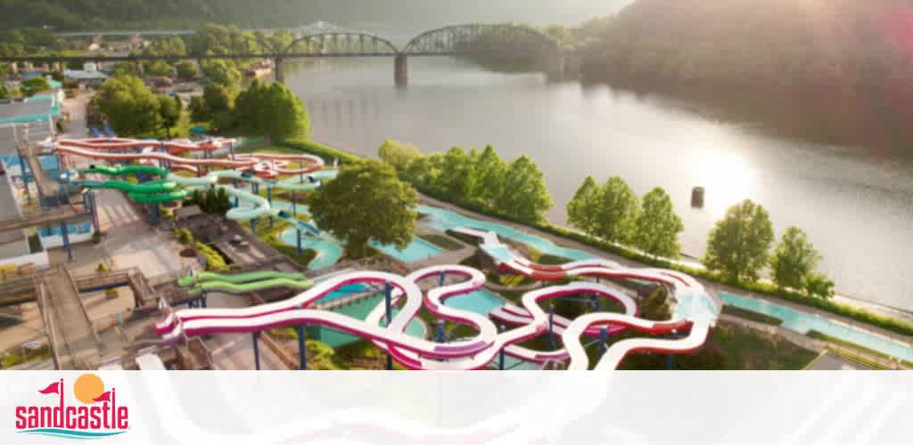 Aerial view of Sandcastle waterpark during a sunny day. Multiple colorful water slides twist and turn above pools of water. In the backdrop, a serene river alongside lush greenery with a bridge crossing it completes the scenic landscape.