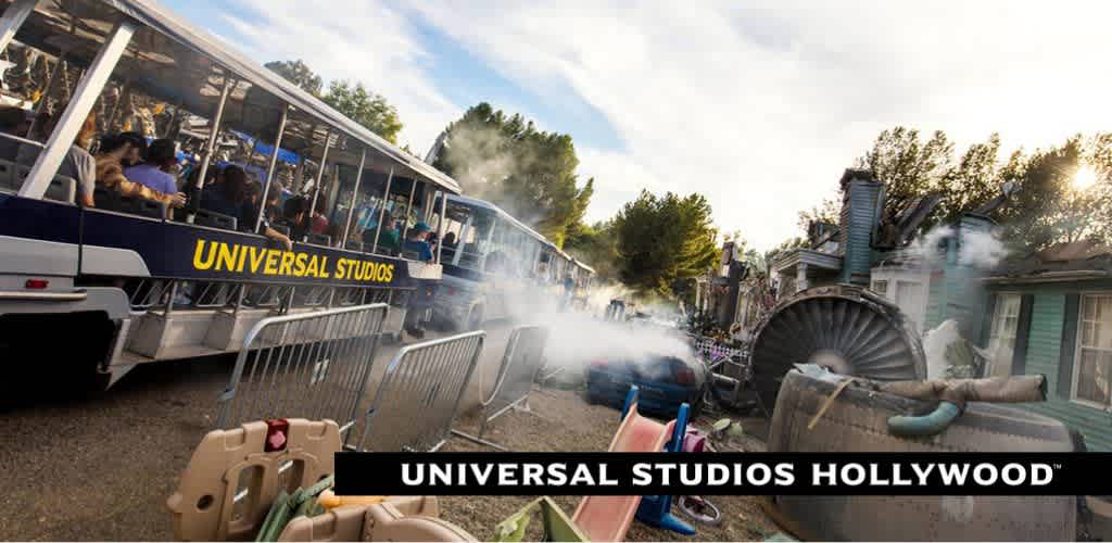 Image shows a tram full of passengers at Universal Studios Hollywood on a studio tour. In the foreground, staged special effects create a scene of chaos with steam and flipped over props, indicating an action-packed adventure. The sky is clear, and the excitement is palpable.