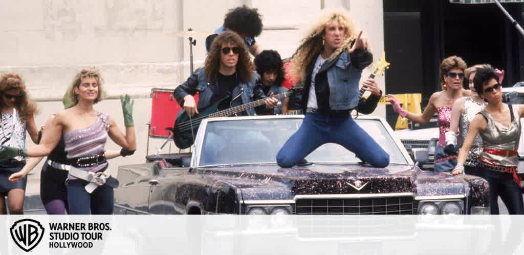 Group of musicians and dancers in 80s attire posing around a vintage car.