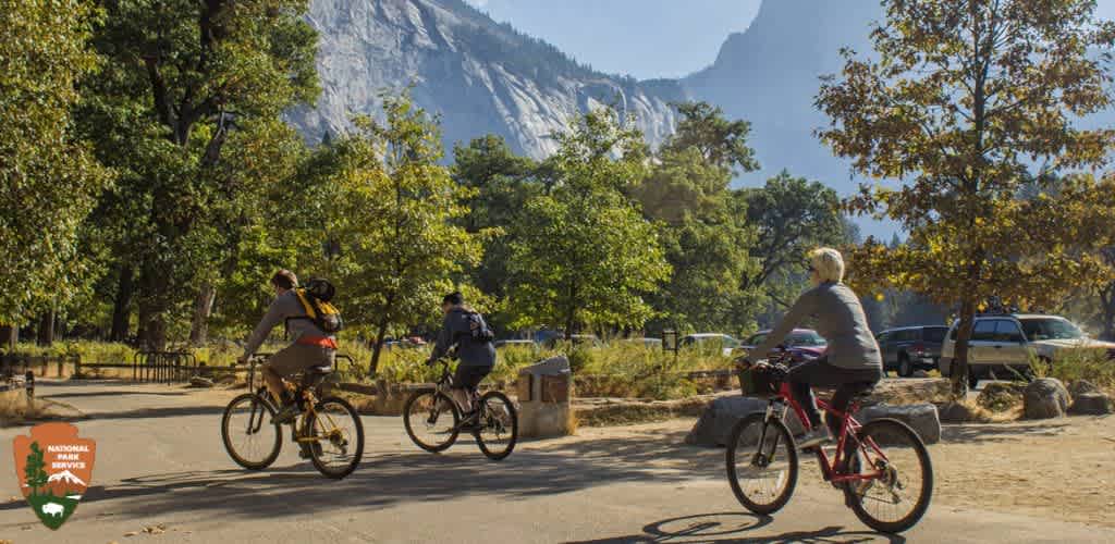 This image shows three individuals riding bicycles on a paved path, likely within a national park setting, given the logo present in the lower left corner suggesting park affiliation. Each cyclist is wearing casual clothing suitable for outdoor activity, and helmets for safety. The bicycles appear to be mountain or hybrid styles, adaptable to the mixed terrain of the park. They are moving away from the viewpoint, heading towards towering cliffs and lush trees signaling the grandeur of their natural surroundings. The landscape is bathed in the warm glow of sunlight, and the clear blue sky suggests favorable weather conditions for outdoor pursuits like cycling.

In the background, vehicles are parked to the right side, indicating proximity to accessible parking areas or roads within the park. The juxtaposition of human activity and nature's majesty encapsulates the allure of outdoor exploration and adventure.

Moreover, for those inspired to embark on similar outdoor adventures or for those looking for exhilarating experiences, GreatWorkPerks.com offers the opportunity for substantial savings on tickets and passes that lead to lifelong memories without breaking the bank. Discover the lowest prices for your next adventure right here!