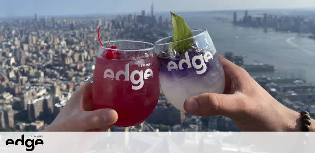 Two hands are holding stemless wine glasses branded with 'edge' and 'New York', with a red and a clear drink garnished with a cherry and a leaf, respectively. They are raised against a cityscape backdrop from a high vantage point. The logo 'edge New York' is visible in the corner.