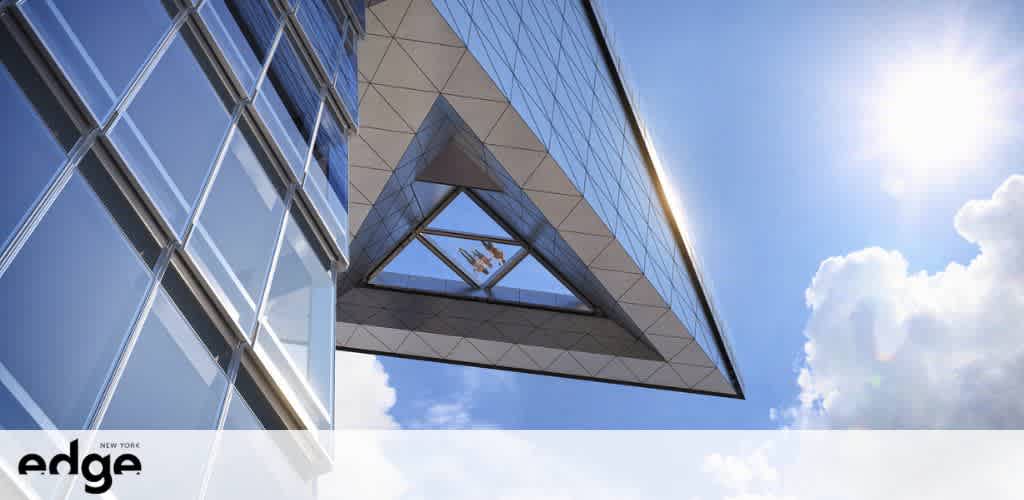 The image shows the exterior of a modern building with a geometric design, featuring a prominent glass facade that reflects the sky. The sun shines brightly from the upper right corner, casting light on the structure and the clouds scattered in the blue sky. The building has a distinct triangular feature that protrudes outward, adding to its architectural uniqueness. The logo of 'edge' with text 'New York' is visible on the lower left side.