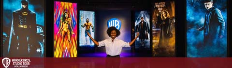 An enthusiastic person with arms outstretched stands center stage at the Warner Bros. Studio Tour. Behind them, a vibrant display features iconic characters from notable Warner Bros. movies. The atmosphere conveys excitement and cinematic fascination.