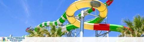 Water slide at Schlitterbahn Waterpark with a bright blue sky in the background. A multicolored closed-tube slide spirals down into a yellow and green open-air slide section with flowing water. Palm trees and signage visible.