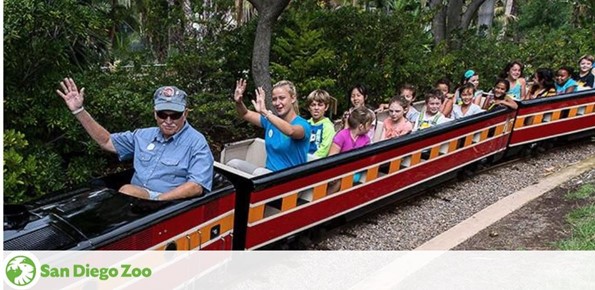 Visitors enjoy a ride on the San Diego Zoo guided bus tour. The image shows a group of smiling passengers with raised hands, expressing excitement. A train conductor in the foreground leads the tour. The setting is lush with greenery.