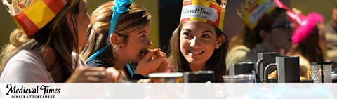 Group of people enjoying a meal at Medieval Times, wearing colorful paper crowns. Smiles indicate a festive atmosphere typical of the themed dinner and tournament experience. The Medieval Times logo is visibly displayed in the lower right.