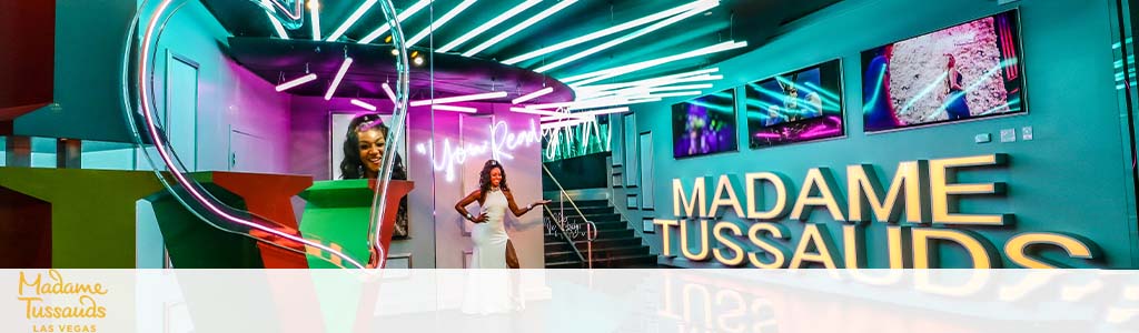 Entrance to Madame Tussauds in Las Vegas featuring vibrant neon lights and a staircase on the right. Two life-like figures are seen posing, with a heart-shaped neon installation to the left. The logo is prominently displayed on the right.