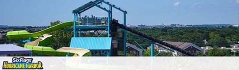 Image shows a tall water slide at Six Flags Hurricane Harbor with a clear sky. The slide has a steep drop and curves, in hues of blue and green. Trees and buildings are visible in the distance, suggesting the park is near a developed area.