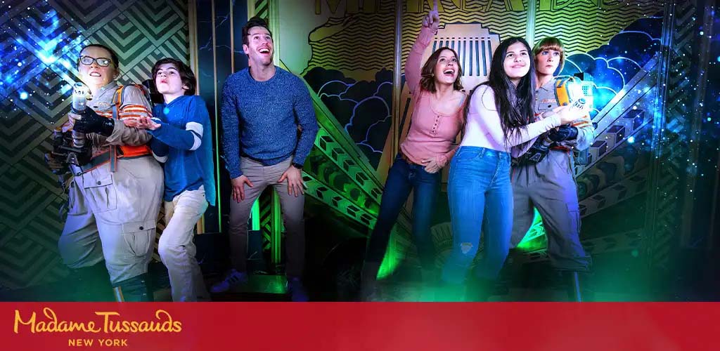 Visitors at Madame Tussauds New York pose excitedly with a wax figure dressed as a Ghostbuster. The figure, equipped with a proton pack, stands to the left while three guests, two women and a man, mimic ghost-busting actions and expressions. Bright, colorful lights enhance the playful atmosphere of this interactive exhibit.