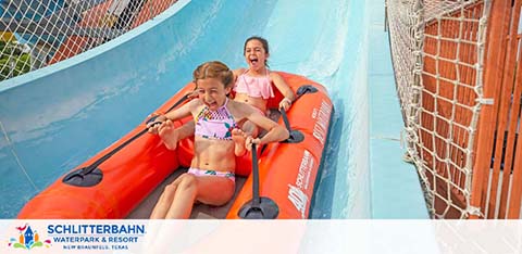 Two people are joyfully sliding down a water slide in an orange raft at Schlitterbahn Waterpark & Resort. They appear to be having a great time, with expressions of excitement and fun. The slide is blue, and safety nets are visible on the side. The waterpark name is prominently displayed at the top of the image.