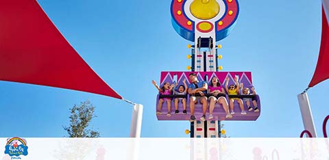 A drop tower ride with excited riders against a blue sky, flanked by red shades.