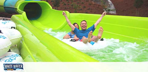 Image featuring two individuals, one behind the other, sliding down a bright green water slide with water splashing around them. Both are smiling with arms raised in excitement. The logo at the bottom reads 'WHITEWATER' indicating a water park setting. They are surrounded by a tunnel-like structure with an open top revealing daylight.