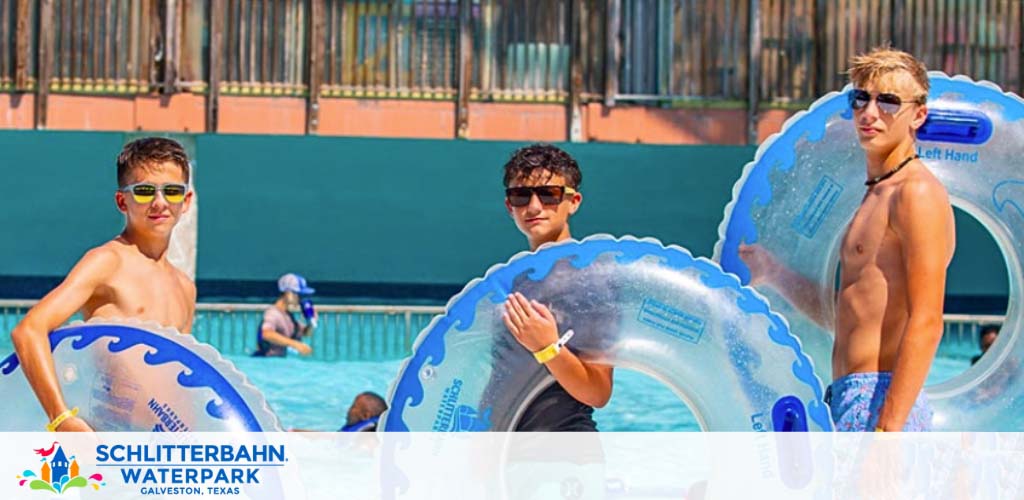 Image shows three young males at Schlitterbahn Waterpark in Galveston, Texas, standing in a pool holding blue inflatable inner tubes, indicative of water-based recreation. The atmosphere is sunny and cheerful, suggesting fun leisure activities.
