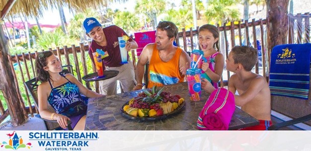 A festive scene at Schlitterbahn Waterpark with a family enjoying refreshments. Two children beam with joy holding water bottles, seated at a table adorned with a fruit platter. A server in a blue cap presents a dish to the smiling adults. Behind, bright towels and park branding embellish the cabana's tropical ambiance.