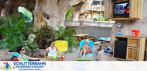 Relaxed visitors enjoy a lounge area at Schlitterbahn Waterpark and Resort. They are seated on colorful chairs amidst tropical decor, with a flat-screen TV on the wall and greenery around. The environment appears to be a cave-like structure with a water park vibe. A red cup and folded towels indicate leisure activities.