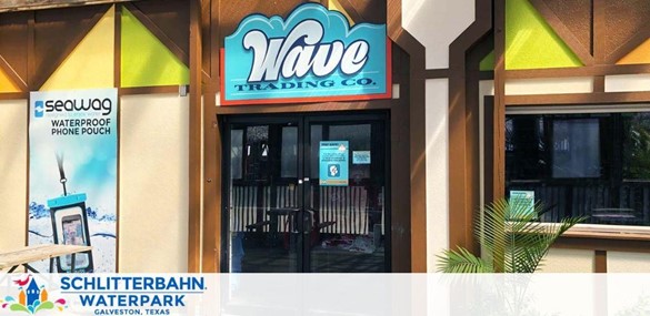 Entrance to Wave Trading Co. at Schlitterbahn Waterpark in Galveston, Texas. The storefront has bright signage, adjacent to banners for waterproof phone pouches, under a sunny sky.