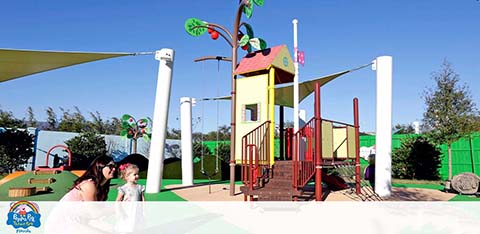 A colorful playground with a slide and climbing structures under shade canopies, with people nearby.