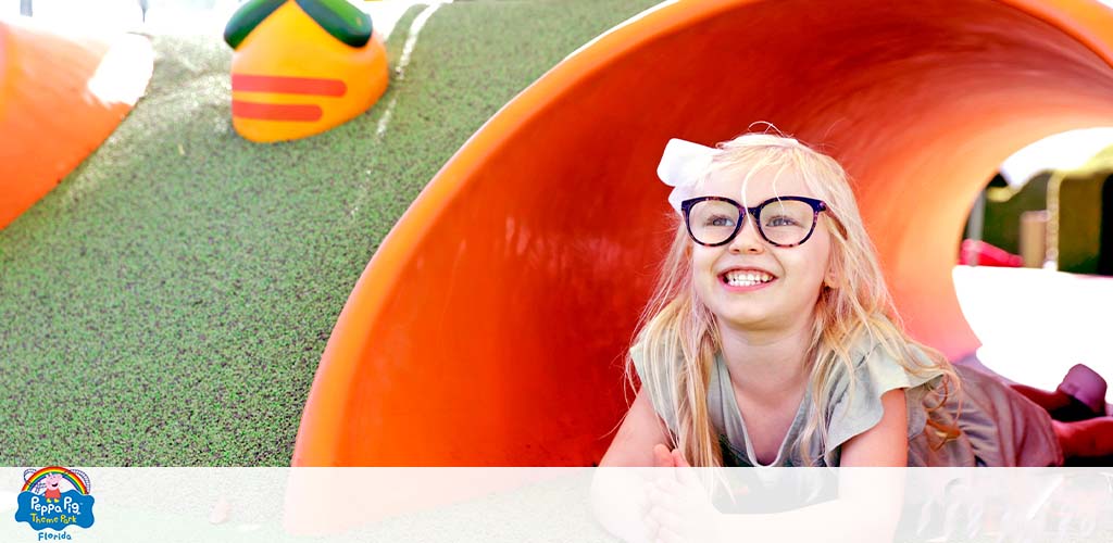 Child with glasses exiting a slide at Peppa Pig Theme Park, smiling brightly.