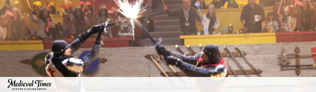 Image shows a medieval-themed jousting event. Two knights in armored costumes with helmets are on the ground clashing with sparks flying from their weapons. A blurred crowd watches in the background, and the logo for Medieval Times Dinner & Tournament is visible.