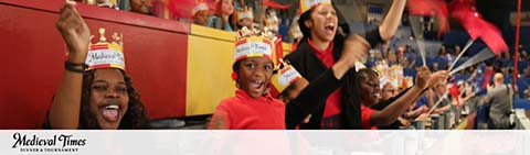 This image showcases a lively scene from Medieval Times Dinner & Tournament. It features three individuals in the audience visibly elated and engaged in the experience. From left to right, there is a young girl with her mouth open as if cheering, wearing a paper crown with the Medieval Times logo. Next to her is a young boy with his arm raised, also donning a paper crown and holding a red flag or pennant. An adult woman stands beside him, also participating enthusiastically in the event, with her mouth open as if shouting and waving a red flag. All three are focusing their attention towards the same point, indicating something exciting is happening out of frame. They appear to be seated in a stadium-like setting with blurred out figures in the background, suggesting a large crowd. The Medieval Times logo is displayed in the bottom left-hand corner. At GreatWorkPerks.com, we believe that unforgettable experiences should be accessible to everyone. That's why we offer discount tickets to amazing events like Medieval Times, ensuring that you can enjoy the excitement at the lowest prices available.