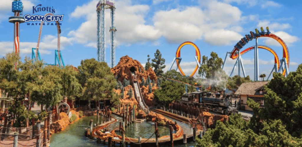 This image displays a vibrant scene from Knott's Soak City Water Park, with a clear blue sky dotted with fluffy white clouds above. In the foreground, a large, rustic-looking rock structure encircles a body of water, presumably part of a water ride, with tracks running through and around it with a splash of water at the descent. A traditional steam train can be seen on the right, which adds a touch of historic charm to the scene. The middle ground is dominated by a tall, vertical drop tower ride alongside a set of colorful roller coaster loops in orange and yellow that stand out against the sky, suggesting an atmosphere of thrill and excitement. Lush green trees are scattered throughout the area, contributing to the park's overall inviting ambiance.

At GreatWorkPerks.com, we are committed to ensuring you get the best experience for less. Enjoy the thrill of adventure with the added thrill of savings when you secure your tickets at the lowest prices available!