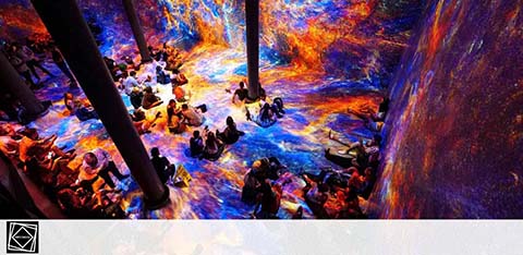 Visitors are immersed in a vibrant, multi-colored space with dynamic projections on the walls and floor, creating an interactive art experience. The room is filled with lights simulating brushstrokes in hues of blue, yellow, and red. People sit or stand, engaging with the captivating display around them.