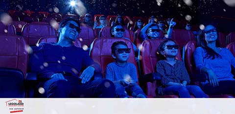 Audience members wearing 3D glasses are enjoying a captivating experience at a LEGOLAND discovery center cinema. Bright light effects suggest on-screen action, highlighting the group's engaged and happy expressions in the dimly lit theater.