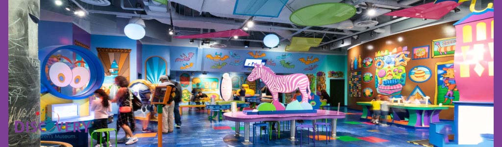 Image shows a vibrant children's museum interior with colorful walls and playful designs. Visitors, including children and adults, are engaged in various interactive exhibits. A large zebra sculpture and tables for activities are visible, creating an inviting and educational environment for families.