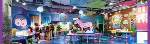 Image shows a vibrant children’s indoor play area with bright colors and whimsical designs. A group of people are interacting in the space, which features a zebra-striped figure, wall decorations with playful characters, and tables for activities. The atmosphere is lively and imaginative.