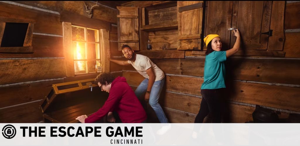 Image shows three individuals in an escape room with rustic wooden walls. One is searching inside an opened trunk, another is looking around the corner, and the third is examining a lock on a door. The logo for 'The Escape Game Cincinnati' is visible in the foreground. Bright light filters through the window, suggesting mystery and adventure.