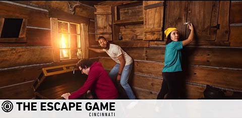 Image shows three people working together in an escape room with rustic wood paneling. One is reaching up to the ceiling, another is examining something intently, and the third appears to be supporting their efforts. The text 'The Escape Game Cincinnati' is displayed at the bottom.