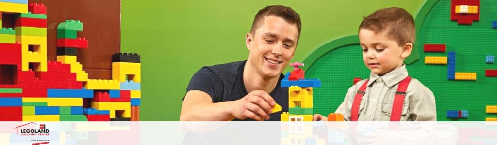 A joyous adult and child engage with colorful Lego blocks at Legoland. The adult, on the left, smiles at the camera, while the child, on the right, focuses on assembling blocks. The background is adorned with vibrant Lego creations, and the Legoland Resort logo is visible.