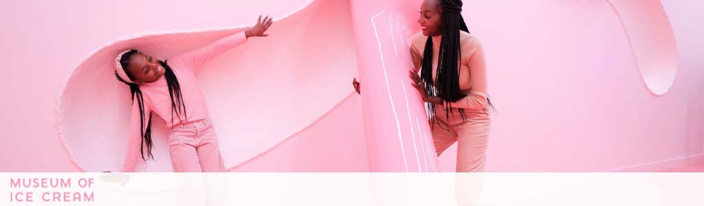 Image shows two individuals in a pink-toned room at the Museum of Ice Cream. One person is playfully interacting with a large, semi-transparent pink sheet, while the other observes with interest. The atmosphere conveys a sense of fun and discovery.