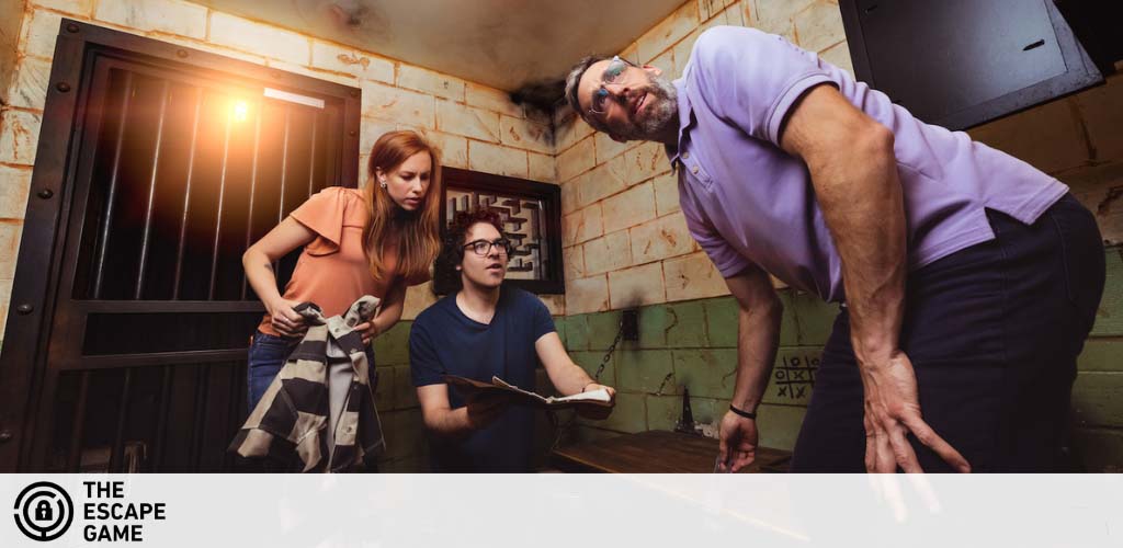 An intense, dimly lit escape room scene. Two individuals analyze a clue, one holding a book, the other wielding a flashlight. A third participant leans eagerly towards a barred window, looking for an escape. They are immersed in the game, solving puzzles together. The Escape Game logo visible, promising adventure.