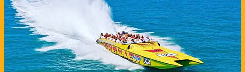A yellow speedboat with passengers creates a large spray on blue water.