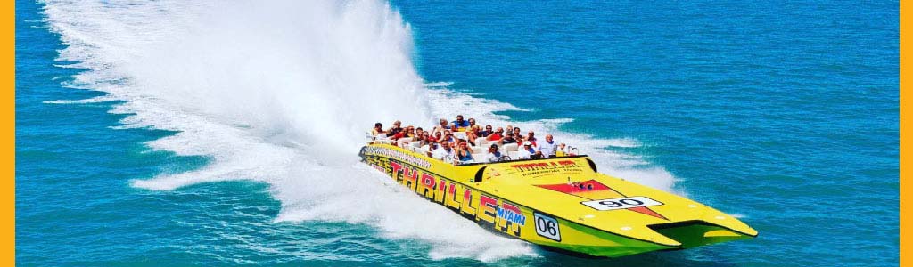 A bright yellow speedboat with passengers creates a large spray on blue water.