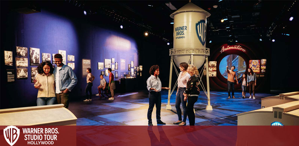 Visitors explore the Warner Bros. Studio Tour Hollywood, viewing exhibits and interactive displays. A couple studies photos, while others chat near a water tower replica. The ambience is welcoming, with warm lights and animation-themed decor.