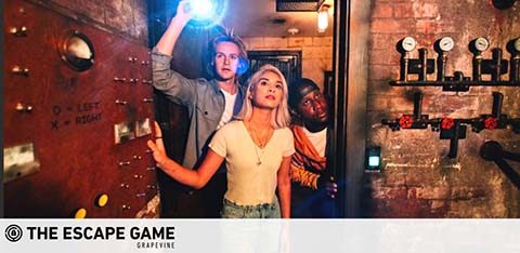 Three friends are engaging in an escape room challenge at The Escape Game Grapevine. They appear focused, with one holding a flashlight above their heads illuminating a room filled with vintage dials and levers, suggesting a puzzle-solving adventure.