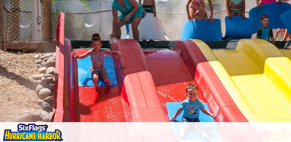 Children enjoy a sunny day at a water park with colorful slides. Two kids slide down red and yellow lanes with water mats, smiling with excitement. In the background, other visitors wait their turn, behind safety fences. The bottom of the image features the Six Flags Hurricane Harbor logo.